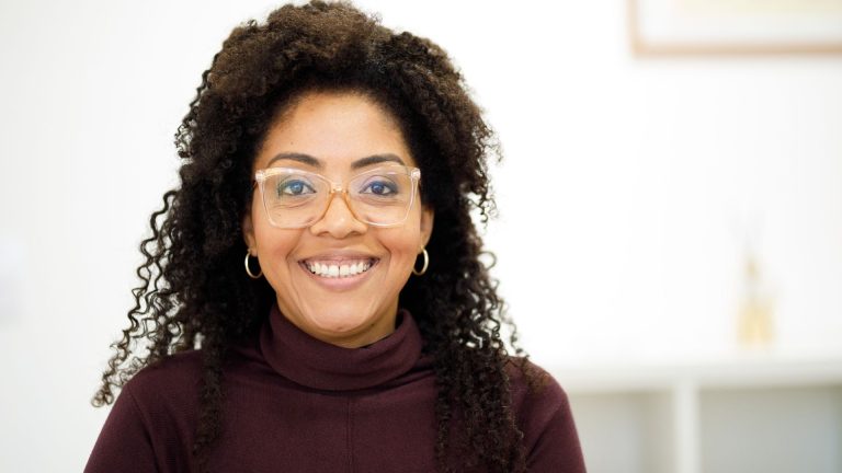 Smiling woman with curly hair wearing glasses and a turtleneck, representing the concept of high-masking autism in adults.