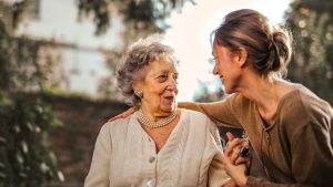 Can Autism Develop Later in Life An elderly woman with short gray hair and pearl jewelry smiles warmly at a younger woman with brown hair tied in a bun, who is holding her hand and engaging in conversation outdoors. The image represents the question Can Autism develop later in life